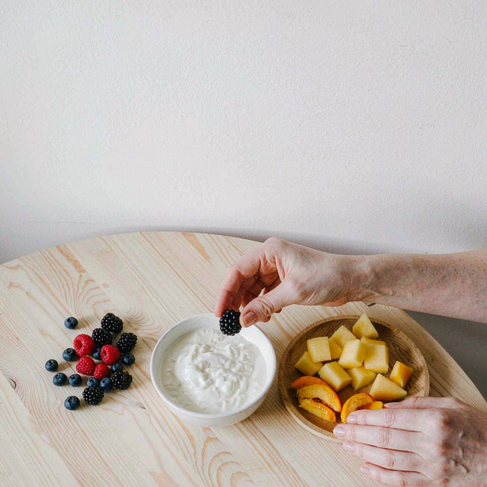 Overhead flat lay of fresh berries and a yogurt bowl arranged on a light table by Caucasian hands with fair light skin