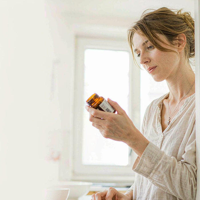 Smiling woman with light brown hair reading an amber supplement jar in a sunlit kitchen