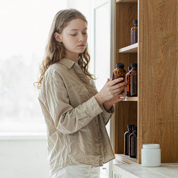 Young woman arranging amber supplement bottles on a bright oak kitchen shelf in soft morning light