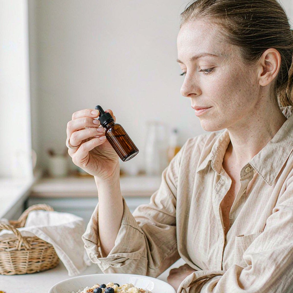 Woman taking an amber dropper supplement beside a bowl of oatmeal breakfast in a bright airy morning kitchen