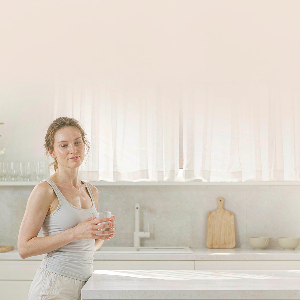 Fair-skinned woman with light brown hair holding a glass of water in a sunlit modern kitchen after her morning workout, bright natural light