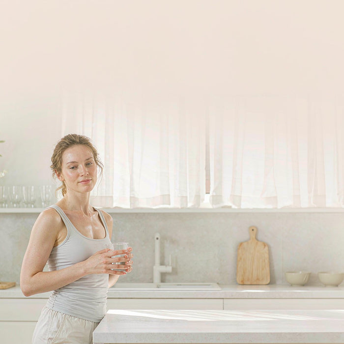 Fair-skinned woman with light brown hair holding a glass of water in a sunlit modern kitchen after her morning workout, bright natural light