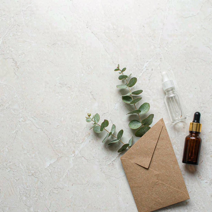 Overhead view of an amber glass dropper bottle and sample vial beside a kraft card and greenery on a pale stone countertop in bright daylight.