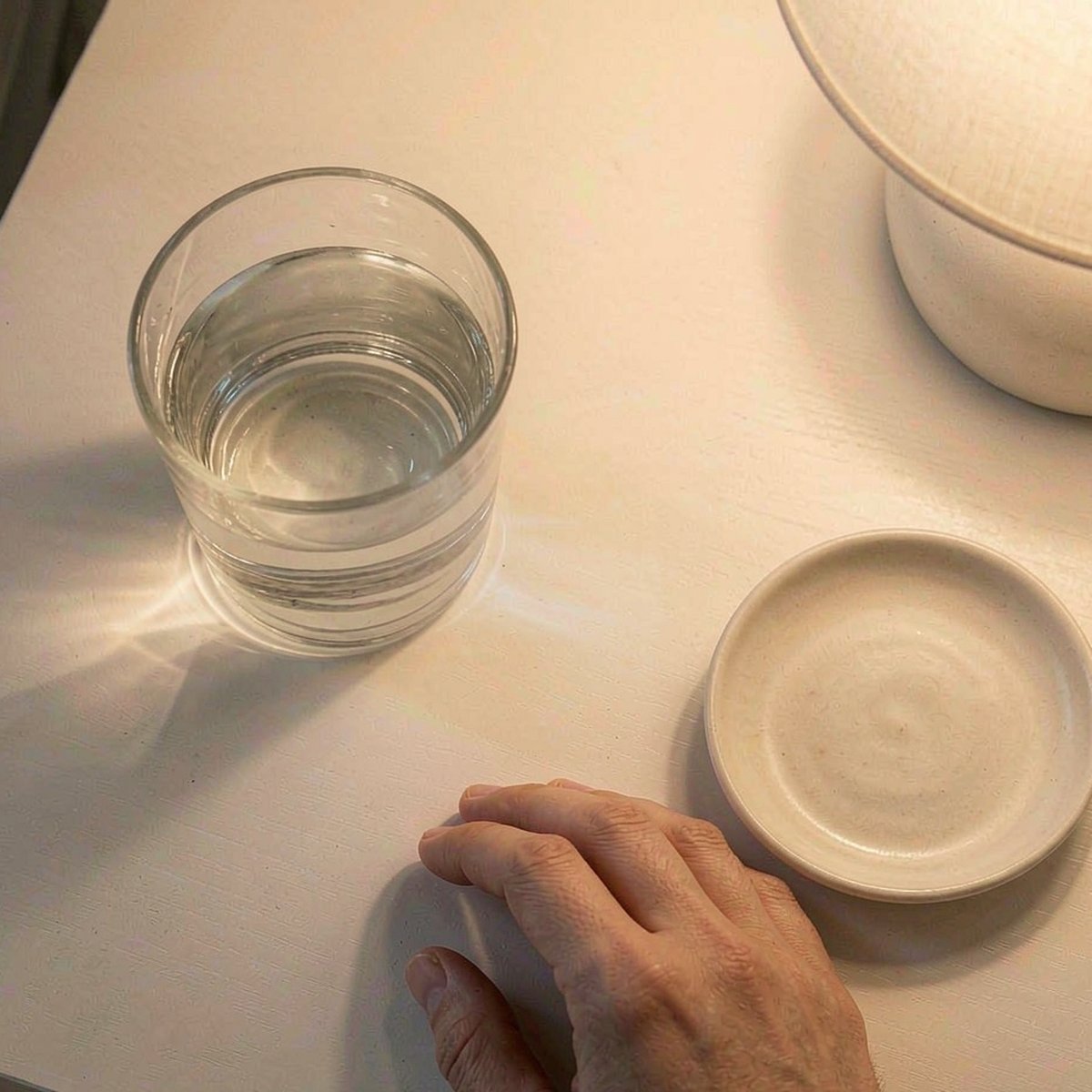 Overhead flat-lay of a minimalist white nightstand with a glass of water and a ceramic dish, warm lamp glow
