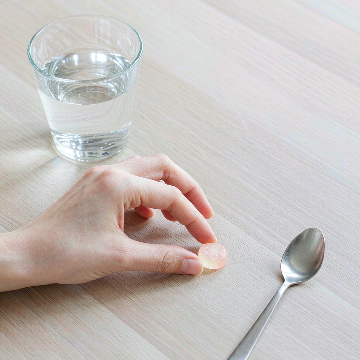 Overhead flat-lay of a fair-skinned hand placing a gummy vitamin beside water and a spoon on a pale oak surface