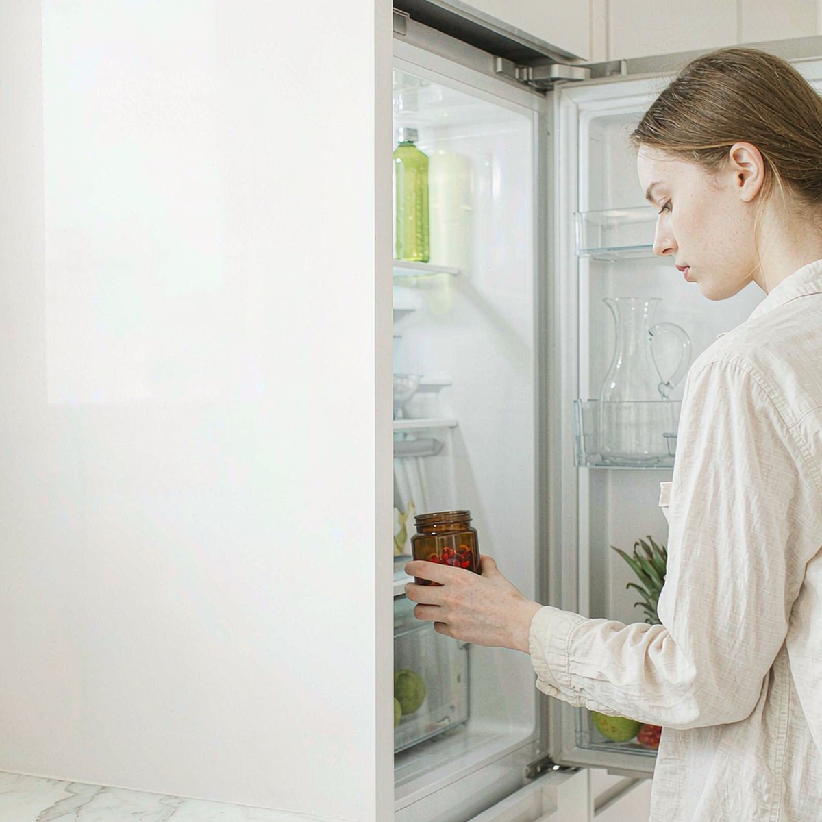 Woman with light brown hair taking an amber gummy jar out of a bright white kitchen refrigerator