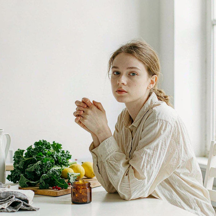 Young woman in a cream linen shirt beside an amber gummy jar with leafy greens, lemon and berries on a bright kitchen counter