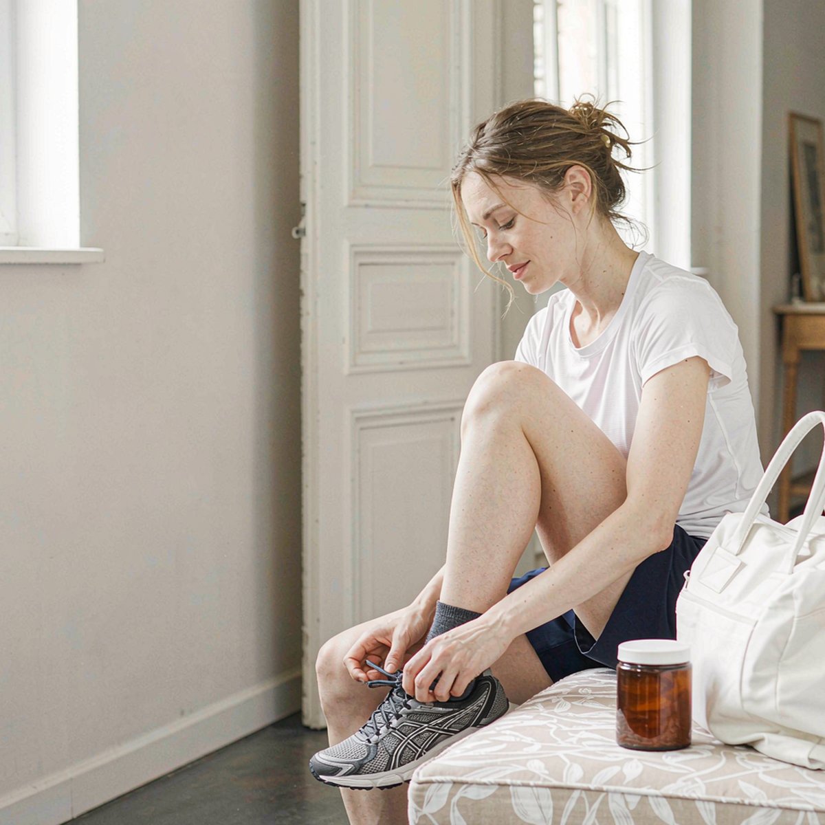 Woman with fair skin tying sneakers beside an amber supplement jar and gym bag in bright morning light