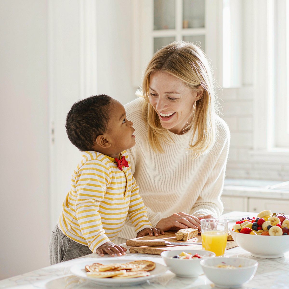 A Caucasian mother with fair light skin smiles while preparing a healthy breakfast for her child in a bright sunlit kitchen