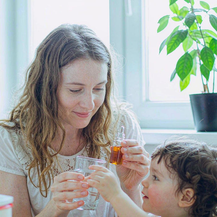 Caucasian mother with fair light skin offering a small herbal dropper bottle with water to her child at a sunlit family table