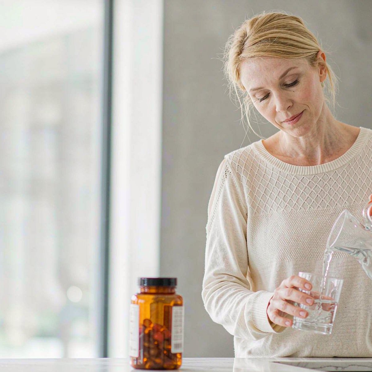 Western fair-skinned woman in a cream sweater pouring water at a bright kitchen counter with a gummy jar