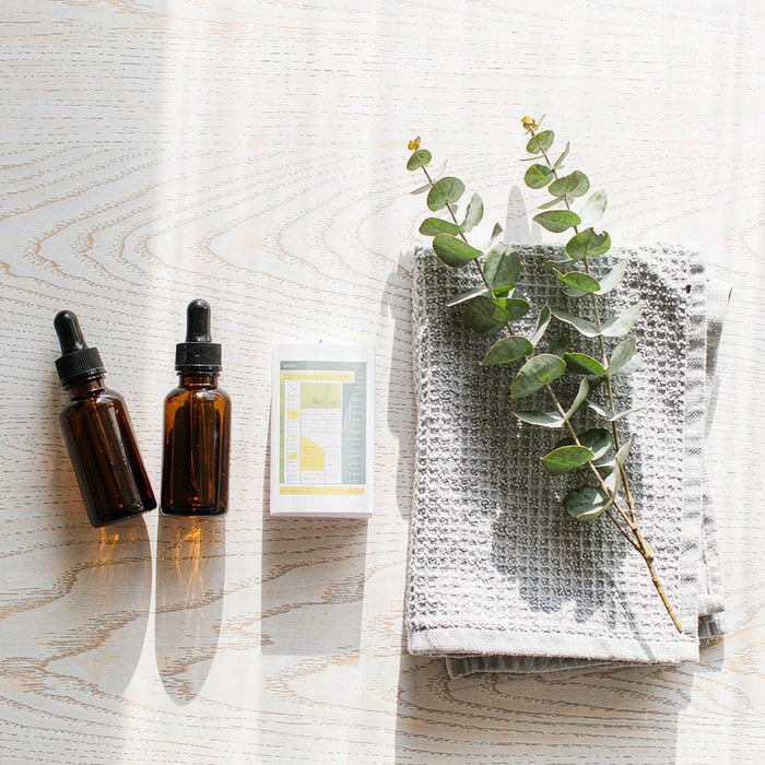 Two amber glass dropper bottles on a white oak table with a blank comparison card, gym towel and eucalyptus sprig in bright morning light