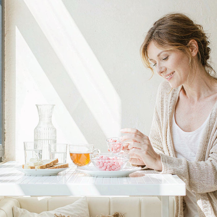 Relaxed fair-skinned woman in her forties holding heart-shaped gummy supplements beside tea on a sunlit breakfast table.