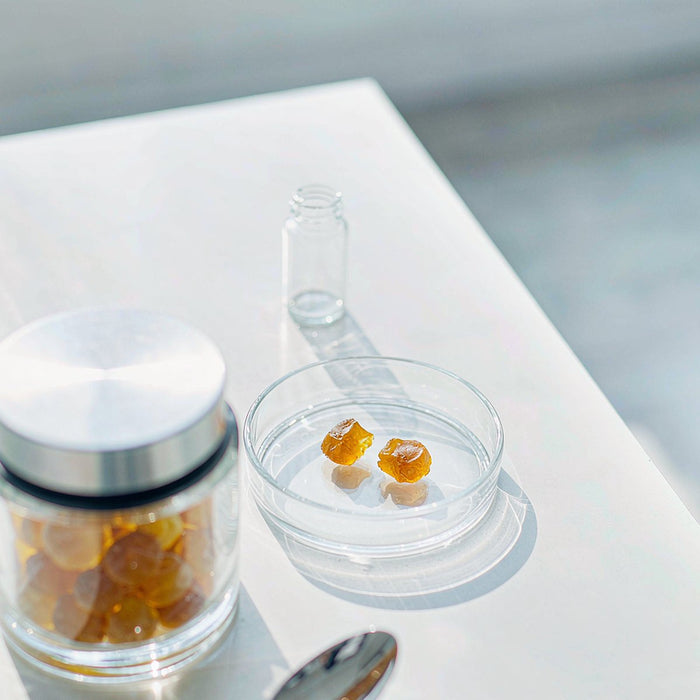 Overhead view of an amber jar of golden gummies with a glass petri dish and sample vial on a bright white lab bench in morning light