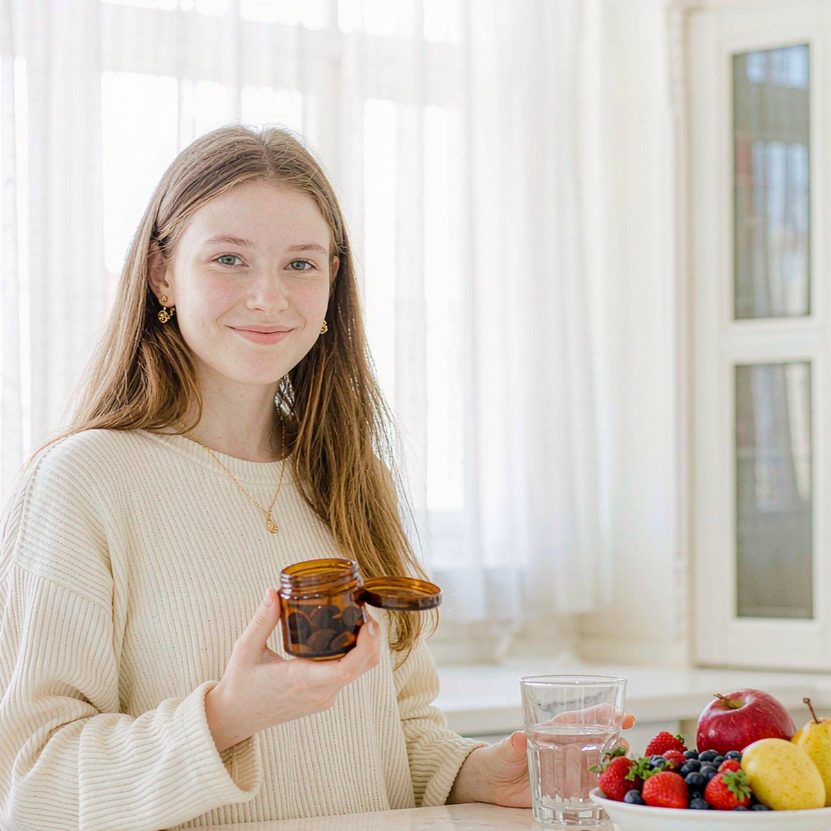 Young woman with light brown hair holding an amber jar of berry gummies in a sunlit morning kitchen