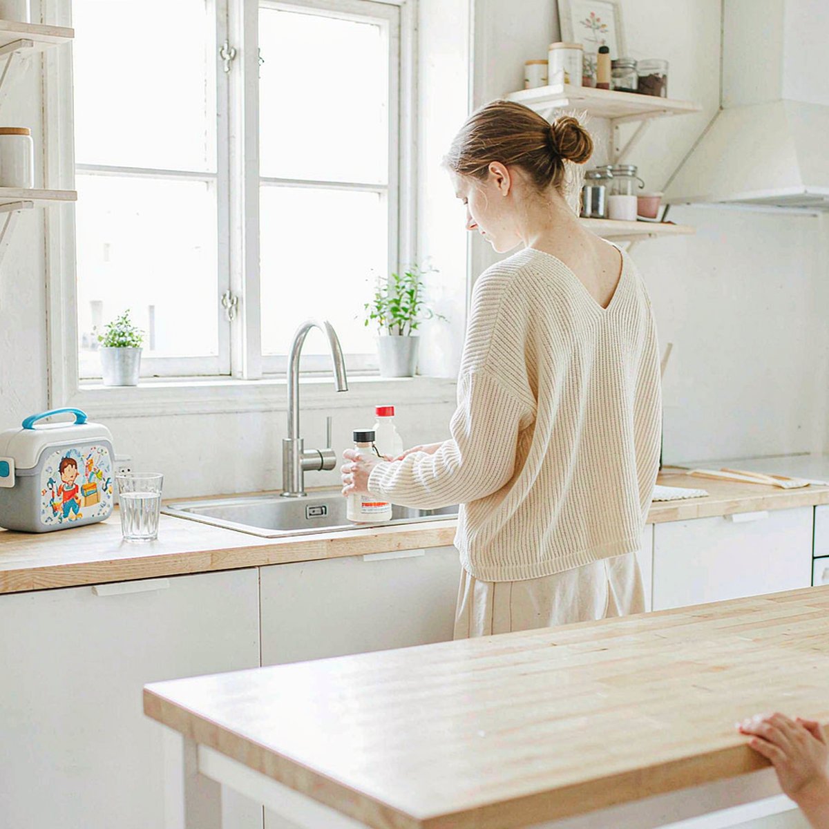 Caucasian mother with fair light skin arranging amber supplement bottles on a sunlit kitchen counter beside a lunchbox.