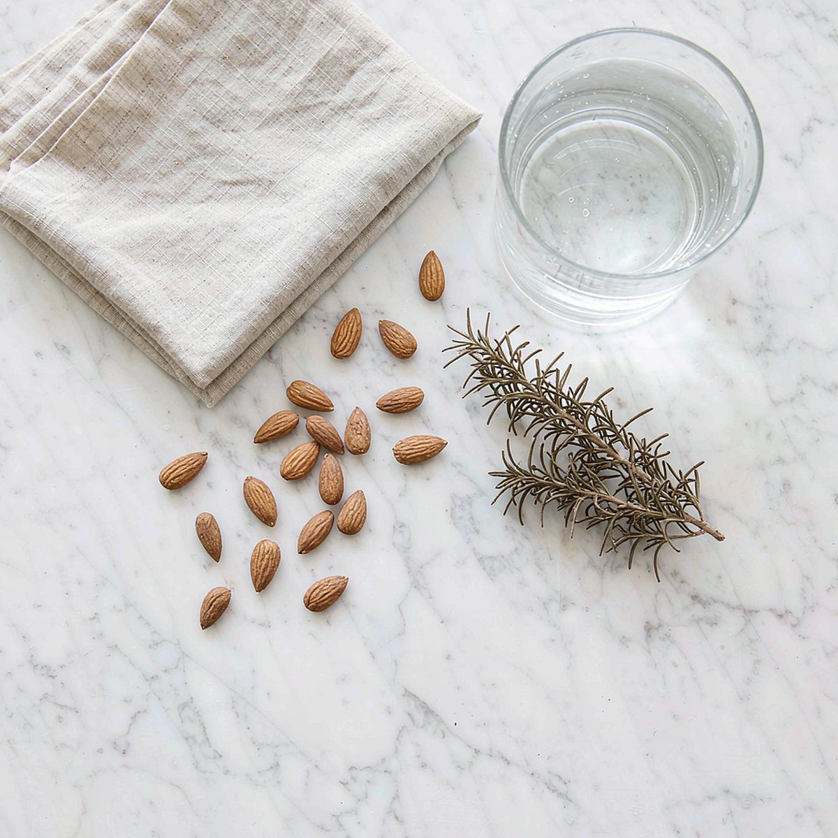 Overhead flat lay on white marble with a glass of water, fresh rosemary, raw almonds and a linen napkin, bright minimalist natural light