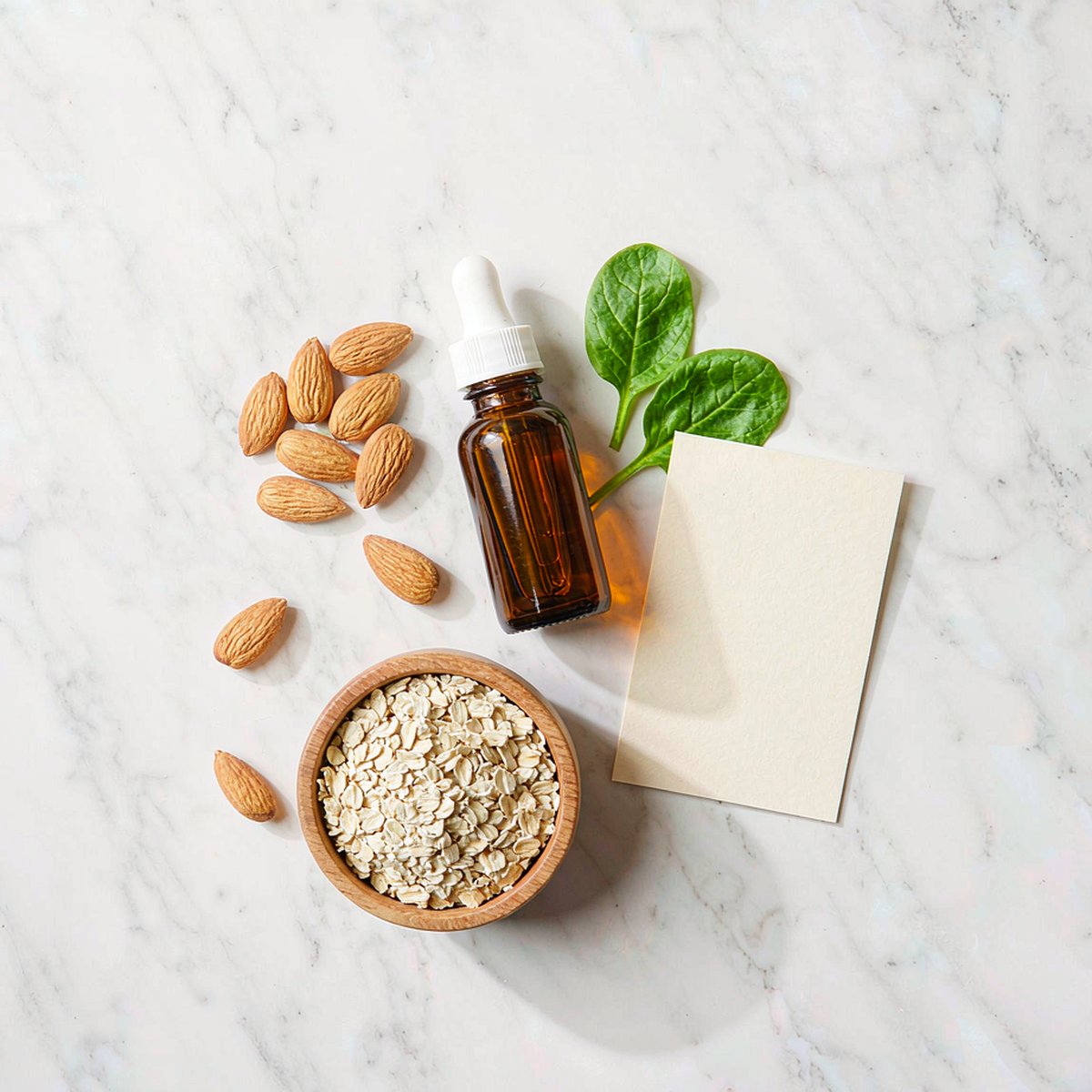 Overhead flat-lay of an amber dropper bottle with almonds, spinach and oats on white marble in morning light
