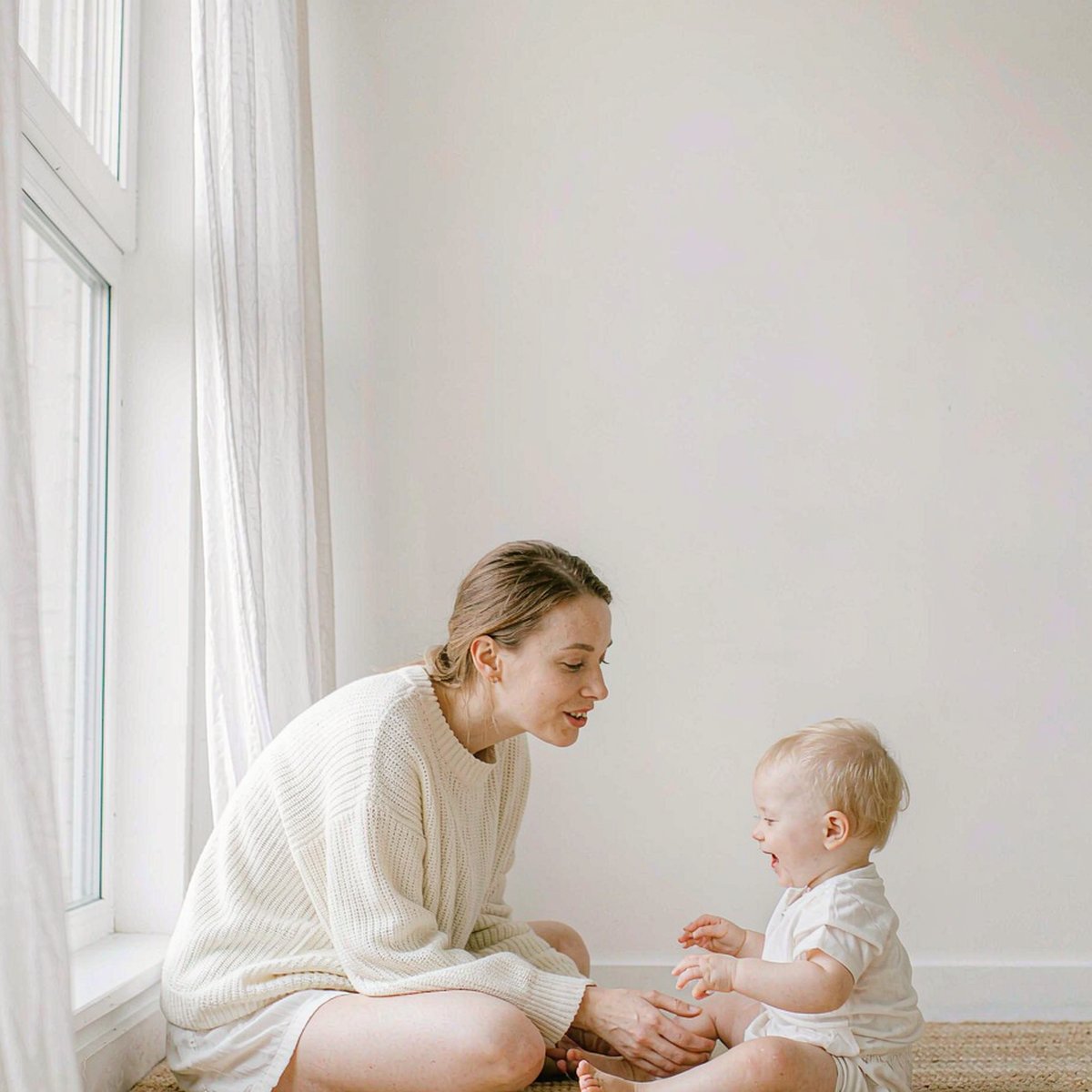 Caucasian mother with fair light skin beside a smiling toddler on a bright minimalist rug, soft daylight