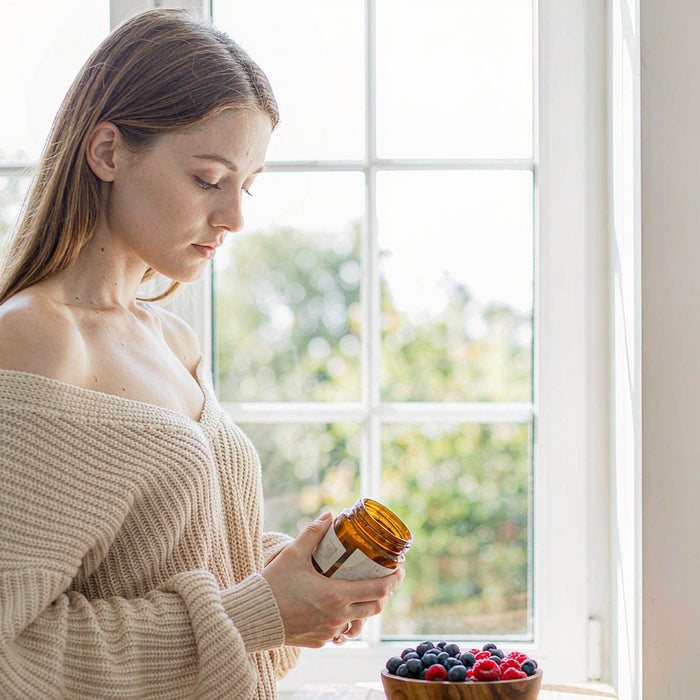 Woman with fair skin holding an amber supplement jar in a sunlit kitchen beside fresh berries
