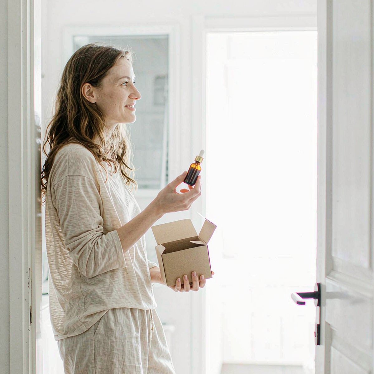 Smiling fair-skinned woman in a bright kitchen doorway holding a kraft delivery box and an amber dropper bottle