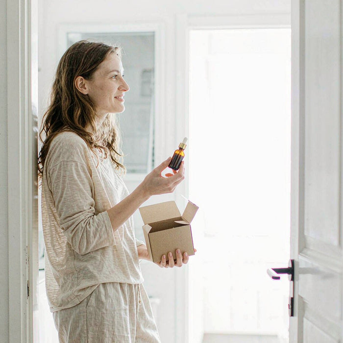 Smiling fair-skinned woman in a bright kitchen doorway holding a kraft delivery box and an amber dropper bottle