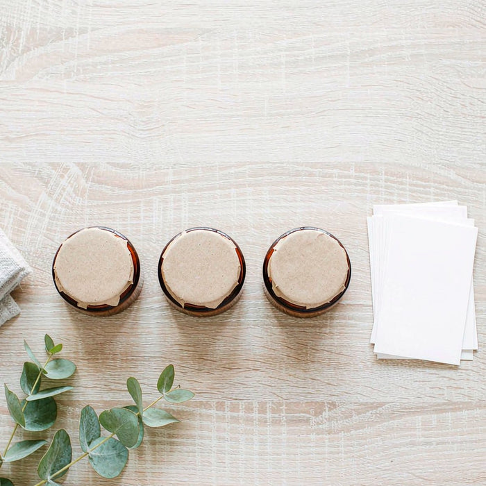 Overhead flat lay of amber jars, eucalyptus sprig and blank cards on pale oak tabletop in bright light